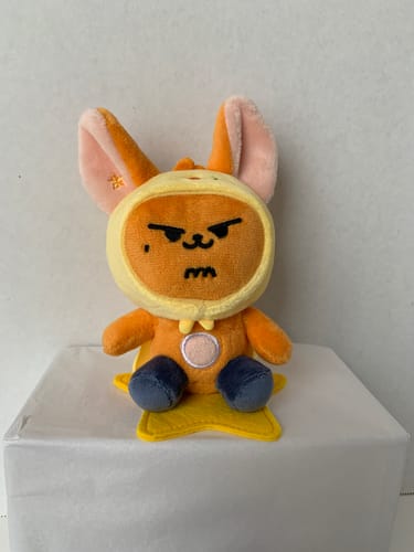 Reviewer's orange TXT PPULBATU plush keyring with an angry face, sitting upright on a white box.