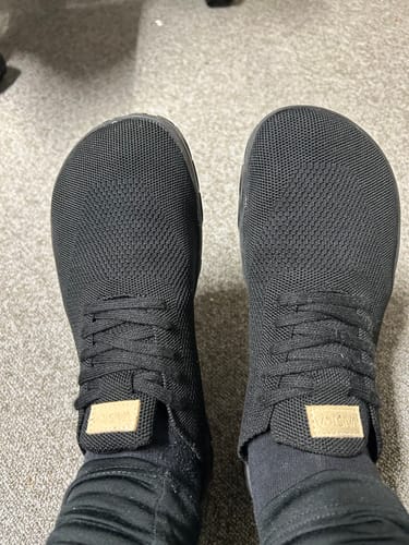 Asiakkaan jalassa olevat mustat Vaistoa Iloa -paljasjalkatennarit ylhäältä kuvattuna.