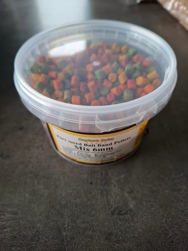Recensent toont een doorzichtige pot met gemengde, gekleurde Flavoured Bait Band Pellets.