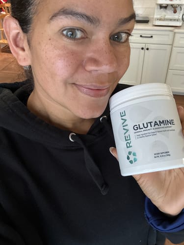 Reviewer holding a white container of Revive Glutamine supplement in their kitchen.