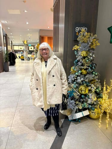 Reviewer smiling while wearing the Beige Faux Fur Double Breasted Longline Jacket indoors next to a Christmas tree.