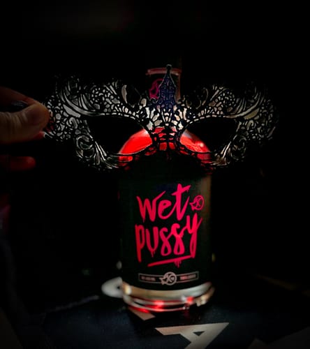Reviewer's bottle of Wet Pussy with glowing liquid, seen behind a detailed mask in a dark room.