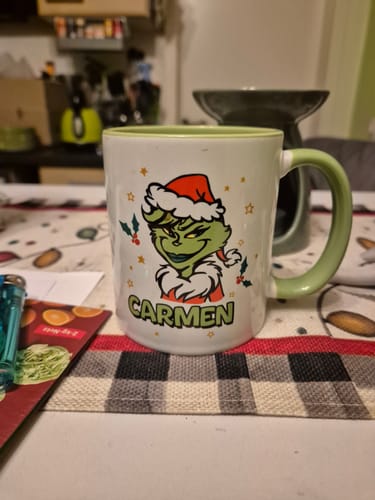 Ein Rezensent zeigt die personalisierte Grinch-Tasse mit dem Namen 'Carmen' auf einem Tisch.
