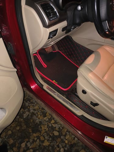 Recenzent pokazuje czarny dywanik samochodowy z czerwoną lamówką, zamontowany po stronie kierowcy.