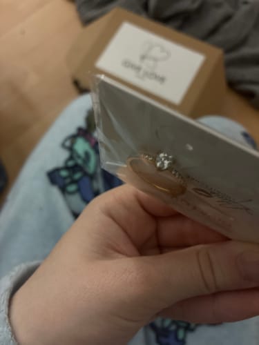 Ein Rezensent hält den Herz Ring in der durchsichtigen Verpackung in der Hand, mit der Versandbox im Hintergrund.