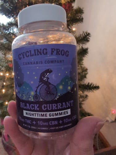 A customer holds a clear bottle of Cycling Frog Black Currant Nighttime Gummies in their hand, with a lit tree in the background.