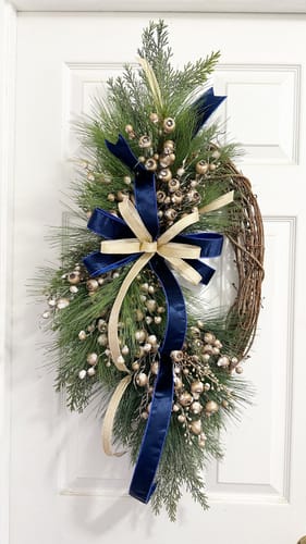 Customer's decorated grapevine wreath hanging on a door with blue ribbons, foliage, and shiny gold berries.