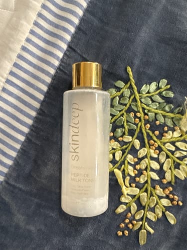 Customer's Dream Glaze Peptide Milk Toner bottle with a shiny gold cap, resting on a dark embroidered surface.