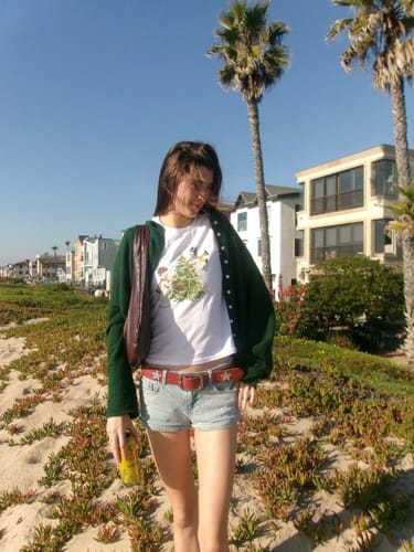 Reviewer on a sandy beach wearing the all that jazz baby tee with a green cardigan and shorts.