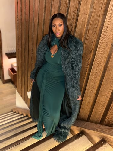 Reviewer wearing the dark green Faux Feather Long Coat over a matching dress while posing on a wooden staircase.