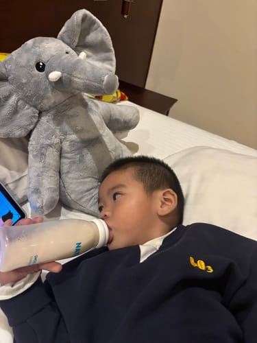 Recensent toont de grijze Olifant verzwaringsknuffel in bed naast een kind dat een flesje drinkt.
