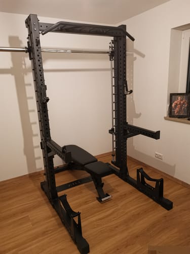 Rezensent zeigt das aufgebaute, schwarze R7 Saber Squat Rack mit Hantelbank in einem Trainingsraum.