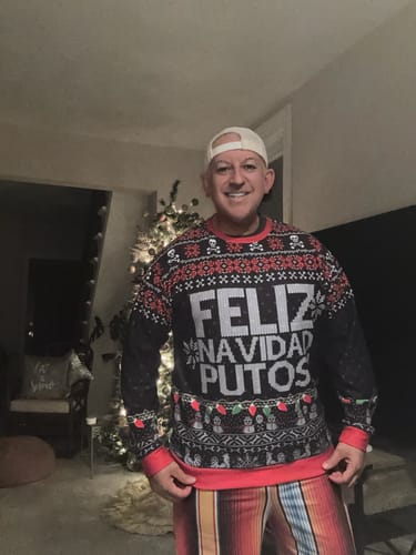 Customer wearing the festive FELIZ NAVIDAD PUTOS Sweatshirt in front of a Christmas tree.