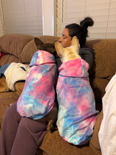 Customer cuddling two dogs wearing matching Velvet Dog Hoodies in the Snow Cone pattern on a couch.