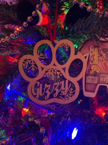 Reviewer's personalized wooden ornament, a paw print with the name "Gizzy," hanging on a Christmas tree with colorful lights.