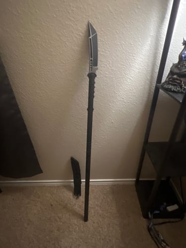 Customer's M48 Sabotage Tactical Survival Spear standing against a wall, with its sheath on the floor beside it.