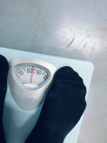 レビュー投稿者がアナログ体重計に立っている様子。針は約83kgを指しており、マンジャロ使用前の体重を示しています。