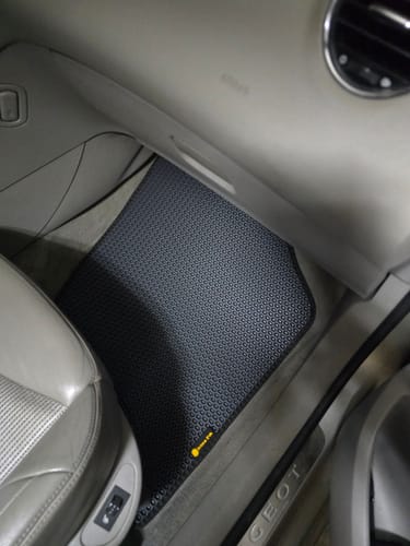 Recenzent pokazuje ciemnoszary dywanik samochodowy z wzorem, idealnie dopasowany do podłogi po stronie pasażera.