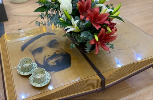 Customer's Wisdom Table, shaped like an open book with an engraved portrait under its glass top, holding a vase and cups.