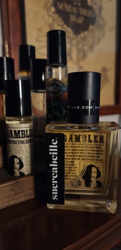 Customer's Gambler perfume in a square bottle and a smaller rollerball on a wooden surface with other bottles.