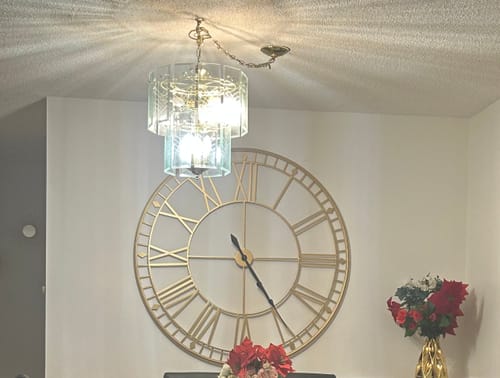 Customer's large gold Roman numeral wall clock mounted on a white wall below a chandelier.