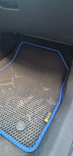 Zbliżenie na czarny dywanik samochodowy klienta z niebieskim obszyciem, położony na podłodze po stronie pasażera.