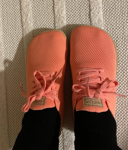 Asiakkaan jalassa olevat korallinväriset Vaistoa Iloa -paljasjalkatennarit mustien sukkien kanssa, ylhäältä kuvattuna.