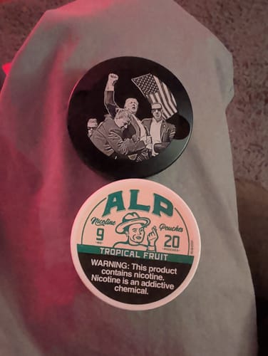 Reviewer's can of Alp Tropical Fruit Nicotine Pouches placed on a gray surface next to another can.