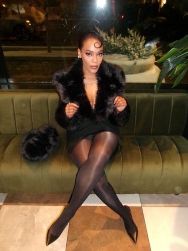 Reviewer on a couch in the Faux Fur Suited Blazer, holding one of the detachable plush fur cuffs.