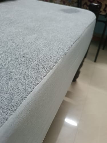 Close-up of the reviewer's gray Flo Mattress Protector, showing the terry cloth texture and fitted corner on a bed.