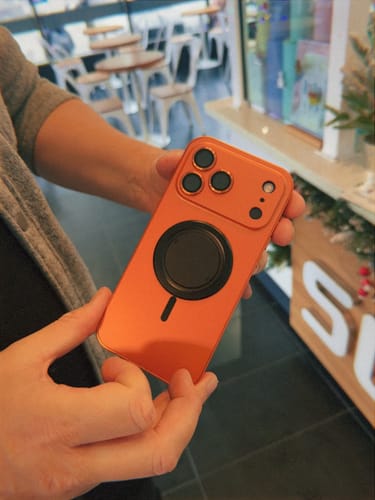 Customer holding their iPhone in the bright orange MagCase Metal, showing the MagSafe ring and raised camera lenses.