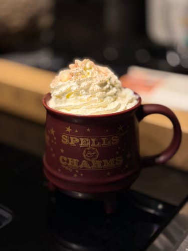 Rezensent zeigt eine burgunderrote 'Spells & Charms'-Tasse, gefüllt mit einem Getränk und Schlagsahne.