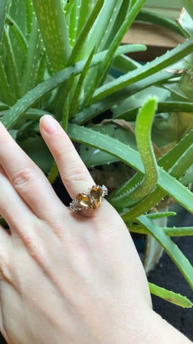 Reviewer's hand wearing the Promise Ring, featuring two coffin-shaped yellow stones, in front of a green plant.