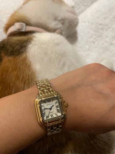 Close-up of the customer's gold GREALY DE MAINS watch, showing its square face and link band.