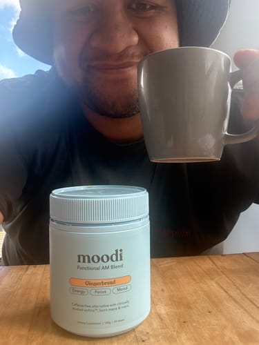Customer smiling and holding a mug, with a container of Moodi Gingerbread blend on a wooden table.
