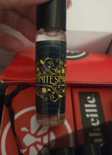 Close-up of a reviewer holding a small rollerball bottle of Infinitesimal perfume with a dark, ornate label.