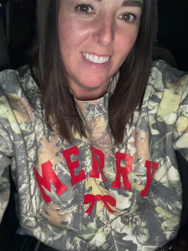 Reviewer smiling in a close-up, wearing the Merry Bow Camo Pullover with its red graphic.