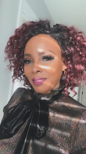 Reviewer smiling while wearing the Janet Collection DEJA wig, which has curly, red velvet-colored hair.