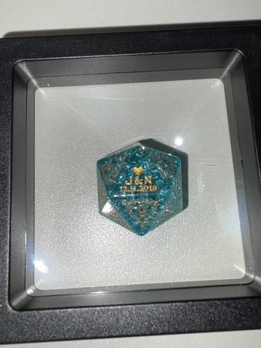 Customer's custom anniversary D20 dice, a sparkly teal die with gold engraving, in a display case.