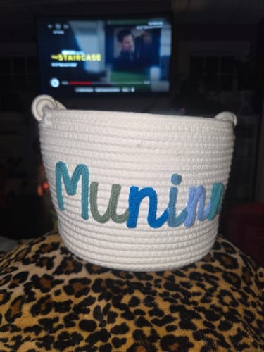 Customer's white personalized knitted basket with the name 'Munim' in multicolor yarn, sitting on a leopard-print surface.
