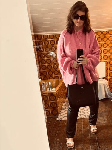 Kundin macht ein Spiegelselfie und trägt den übergroßen pinken Cardigan "CozyGlam" zu einer schwarzen Hose.