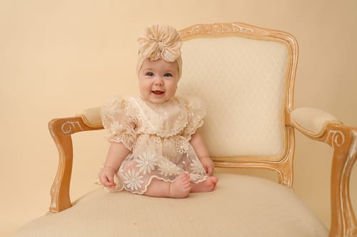 Customer's baby sitting in a chair, smiling while wearing a lacy cream dress and matching bow headband.