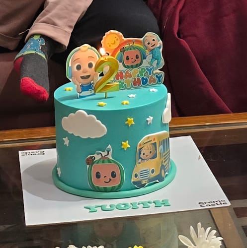 Customer's light blue Cocomelon Song Cake decorated with characters, clouds, and a lit number '2' birthday candle.