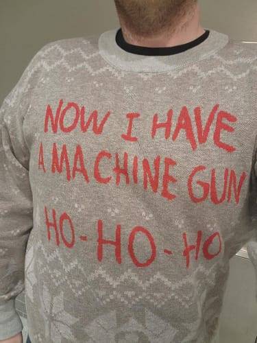 Reviewer wearing the gray knitted Die Hard sweater with the red 'Now I Have A Machine Gun Ho-Ho-Ho' text.