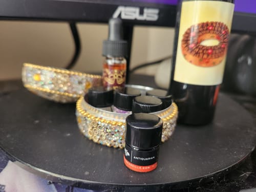 Customer's small sample bottle of Antiquarian perfume in focus on a desk, with other scents in a decorative jeweled container behind it.