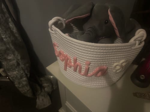 Customer's personalized white knitted basket with 'Sophia' in pink yarn and a flower icon, holding a toy elephant.