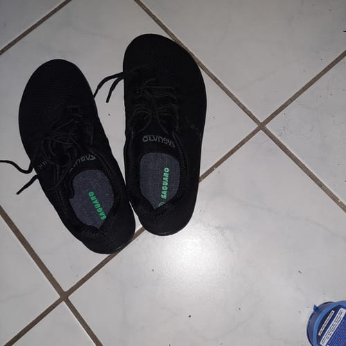 Ein Kunde zeigt ein Paar schwarze Joy II Barfußschuhe von oben auf einem gefliesten Boden.