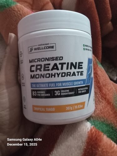 Reviewer holds a white tub of Wellcore Micronised Creatine Monohydrate in Tropical Tango flavor against a blanket.