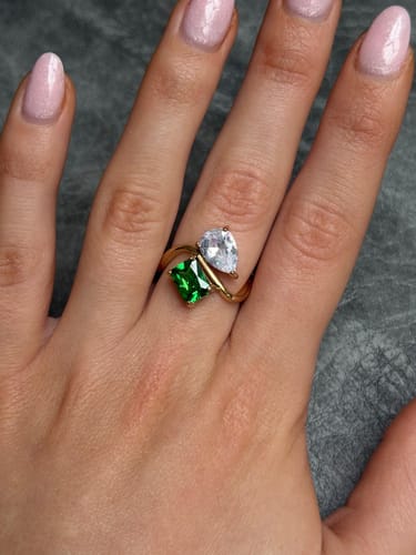 A customer's hand wearing the Navae Birthstone Ring, showing the gold band with a green radiant-cut and a clear pear-cut stone.