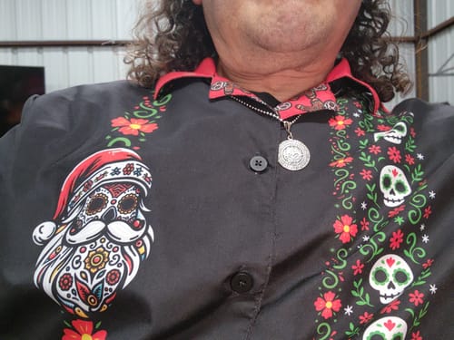 Close-up of a reviewer wearing the black El Sancho Clause Guayabera, showing the printed sugar skull Santa graphic on the chest.
